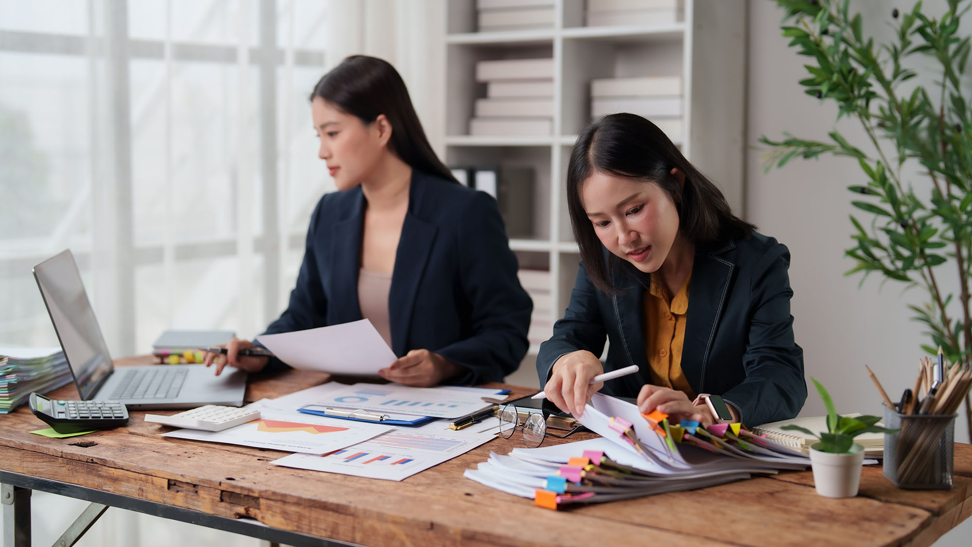 Two professional Asian women working together at a rustic wooden desk, one focused on sorting a stack of papers with colorful binder clips and the other using a laptop for business tasks